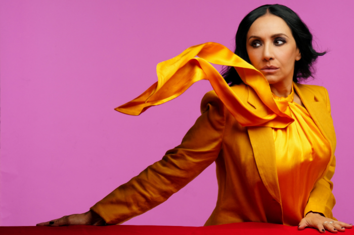 Musician Roxana Amed in a yellow blazer and shirt, looking to the left, with a bright pink background