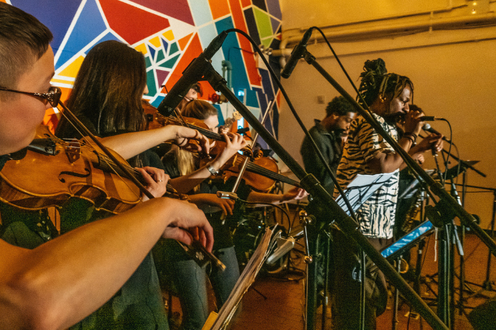 ShoutHouse performing with Spiritchild, geometric shapes form a back drop, several musicians hold string instruments