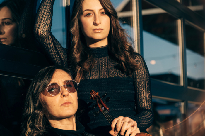 Talla Rouge duo pose with instruments outside in front of windows wearing black dress clothes. One member wears sunglasses.